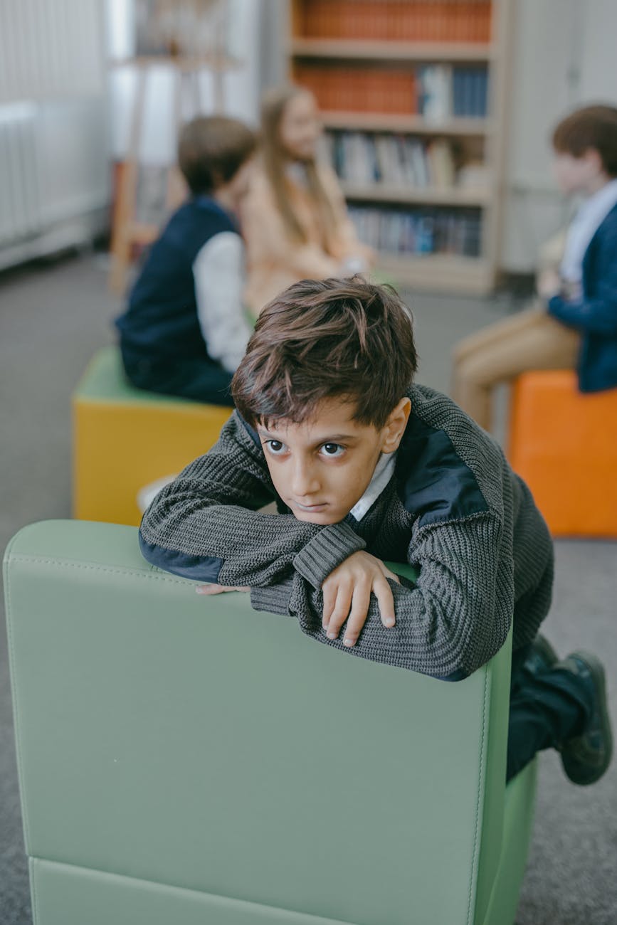 A shy child sitting alone on a school chair looking thoughtful and withdrawn while other kids play in the background, showing social hesitation at school.